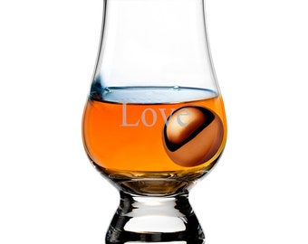 May include: A clear glass whiskey tasting glass with amber liquid and a brown sphere inside. The word "Love" is etched on the glass. The glass has a wide bowl and a short, sturdy stem.