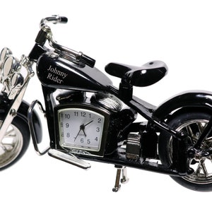 May include: Black motorcycle clock with a silver face and hands. The motorcycle has a chrome exhaust pipe and wheels. The clock face has the words "Johnny Rider" engraved on the gas tank.