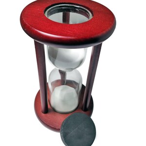 K&ouml;nnte beinhalten: Eine Sanduhr aus Holz mit einem roten Sockel und einer durchsichtigen Glasbirne. Die Sanduhr ist mit wei&szlig;em Sand gef&uuml;llt und hat einen schwarzen Sockel.