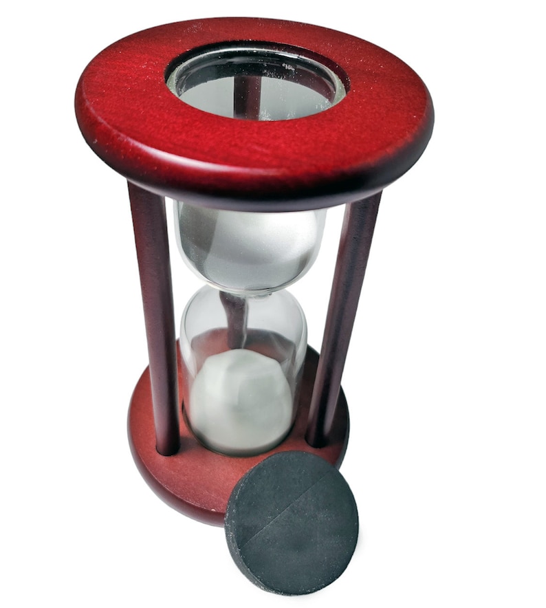 K&ouml;nnte beinhalten: Eine Sanduhr aus Holz mit einem roten Sockel und einer durchsichtigen Glasbirne. Die Sanduhr ist mit wei&szlig;em Sand gef&uuml;llt und hat einen schwarzen Sockel.