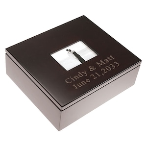 May include: A dark brown wooden box with a photo frame on the lid. The frame shows a black and white image of a couple. The box is engraved with the text "Cindy & Matt June 21, 2033".