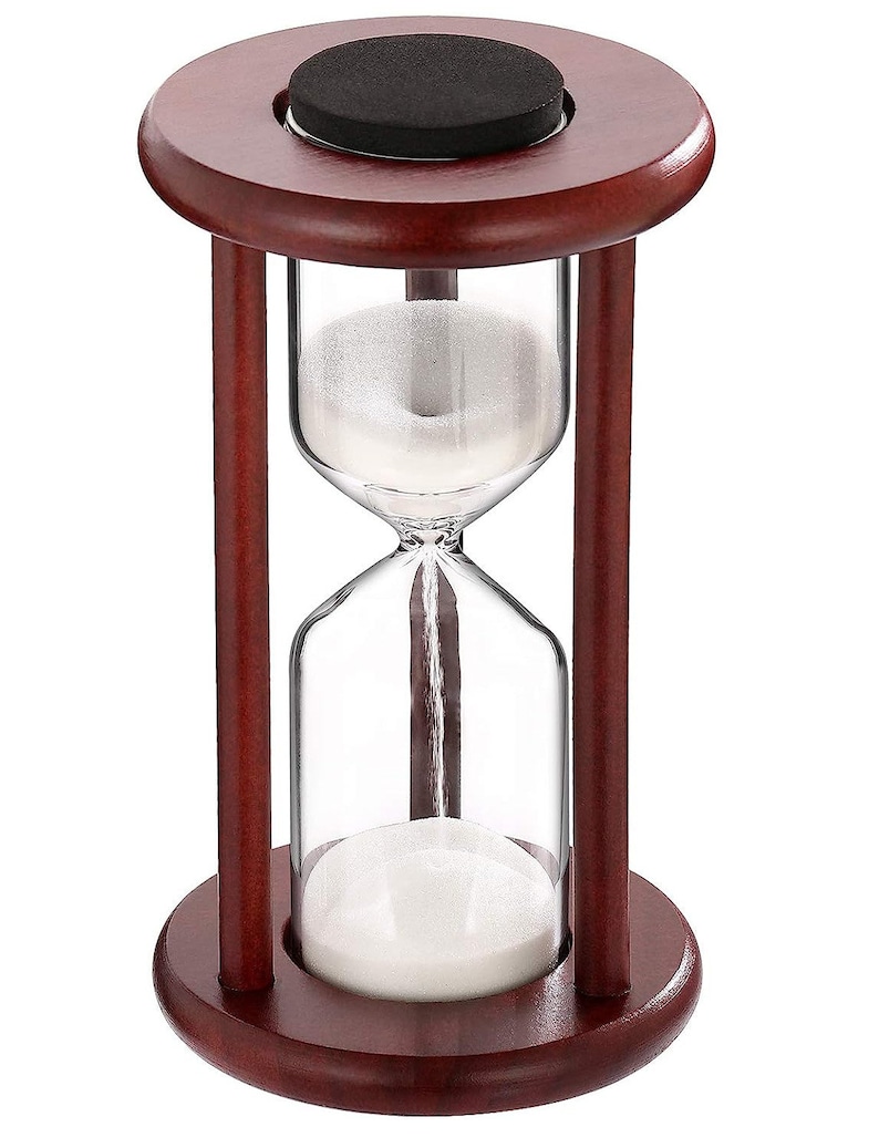 K&ouml;nnte beinhalten: Eine Sanduhr aus Holz mit einer durchsichtigen Glasbirne und wei&szlig;em Sand. Die Sanduhr ist etwa 15 cm hoch und hat einen dunkelbraunen Holzsockel.