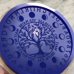 May include: A round, blue stamp with a wooden base. The stamp features a tree design in the center, surrounded by phases of the moon and the words "REPHRASE MAYBE YES WANT ON TIAW" and the alphabet. A unique tool for crafting.
