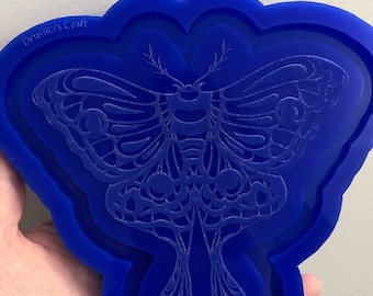 May include: Blue silicone mold with a detailed design of a moth. The mold is shaped like a moth with a raised outline. The text "Drusilla's Craft" is visible in the top left corner of the mold.