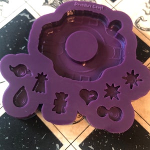 Op de afbeelding: Paarse siliconen mal met een groot cirkelvormig ontwerp en kleinere ontwerpen van een hart, een ster, een druppel, een oneindigheidssymbool en andere abstracte vormen. De mal is gelabeld "Drusillas Craft".