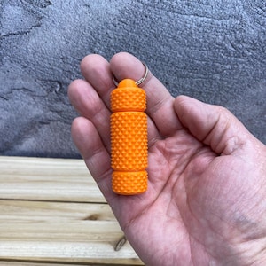 May include: Bright orange, textured keychain accessory. The cylindrical object has a series of raised, conical shapes. The keychain is held in a hand, and the background is a textured gray surface.