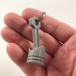 May include: A gray metal keychain shaped like a piston. The keychain has a silver ring for attaching keys.