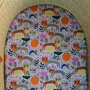 Puede incluir: Una cesta de mimbre tejida con un forro de tela blanca que presenta un patrón repetido de gatos rayados de color naranja, amarillo y negro, flores rosas y hojas verdes.