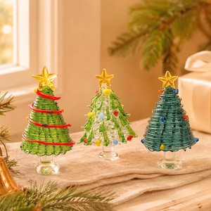May include: Three glass Christmas tree figurines standing on clear circular bases. Each tree is topped with a yellow star and features colorful glass accents, including red ribbons on the left tree and small multi-colored dots on the others.