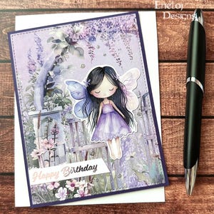 May include: A greeting card with a purple border and a fairy illustration. The fairy has black hair, a purple dress, and blue wings. The card has the words "Happy Birthday" on a white banner. A black pen is on the right side of the card.