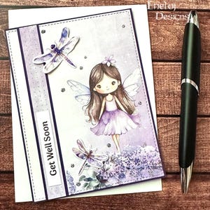 May include: A handmade greeting card with a fairy illustration and the words "Get Well Soon." The card features a purple and white color scheme, with dragonfly accents and sparkling embellishments. A black pen rests on a wooden surface.