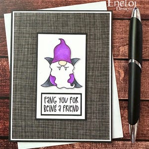 May include: A handmade greeting card featuring a cartoon vampire gnome with a purple hat and cape. The card has the text "FANG YOU FOR BEING A FRIEND" and is set against a textured gray background. A black pen rests beside the card.