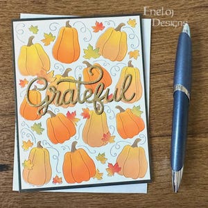 May include: A rectangular greeting card featuring a repeating pattern of orange pumpkins and small autumn leaves on a white background, with the word "Grateful" in gold glitter script across the center, accompanied by a metallic blue ballpoint pen.