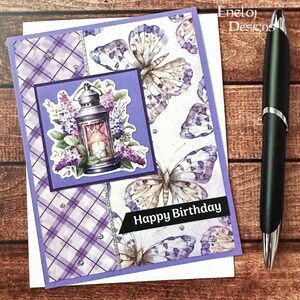 May include: A handmade birthday card with a purple and white butterfly pattern and a square image of a lantern surrounded by flowers. The words "Happy Birthday" are on a black banner. A black pen is on the right.