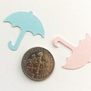 Op de afbeelding: Twee papieren uitsnijdingen van paraplu's, een lichtblauwe en een lichtroze, worden getoond naast een Amerikaanse dime-munt. De paraplu's zijn ongeveer zo groot als de munt.