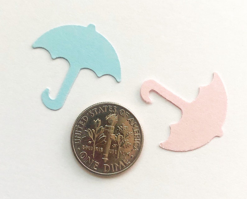 Op de afbeelding: Twee papieren uitsnijdingen van paraplu's, een lichtblauwe en een lichtroze, worden getoond naast een Amerikaanse dime-munt. De paraplu's zijn ongeveer zo groot als de munt.