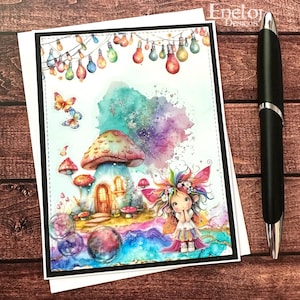 May include: A whimsical greeting card featuring a fairy house with a mushroom roof, colorful lights, and a fairy with a flower crown. The card has a black border and is paired with a black pen on a wooden surface. The card is from Enelor Designs.