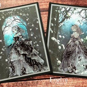 May include: Two art prints featuring a woman in a dark gown standing in a forest setting. The prints have a black border and the artist's name, "Eneloj Designs," is in the top left and bottom right corners. The woman is surrounded by white flowers and trees.