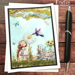 May include: A greeting card with a whimsical illustration of a girl under a daisy umbrella, with a hummingbird and butterflies. A picnic basket and sunflowers are in the background. The card has a black border and is next to a black pen.