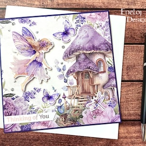 May include: A greeting card featuring a whimsical fairy scene with a mushroom house, butterflies, and a fairy. The card is predominantly purple and white, with the text "Thinking of You" on a banner. A black pen rests beside the card.