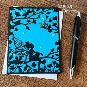 May include: A blue card with a black silhouette of a fairy sitting on a branch with a butterfly flying above. The card is decorated with green glitter.