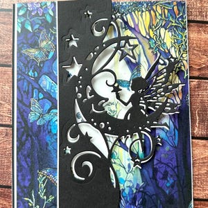 May include: A handmade greeting card featuring a black die-cut design of a fairy on a crescent moon, with stars and swirls. The background showcases a colorful forest scene with butterflies in shades of blue, purple, and green. The card is on a wooden surface.