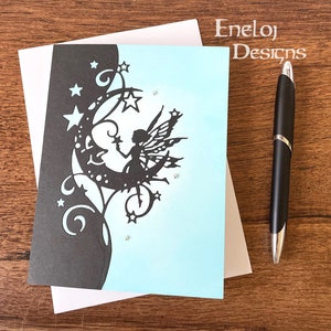 May include: A light blue card with a black silhouette of a fairy sitting on a crescent moon. The fairy is holding a star and has wings. The moon is surrounded by stars and swirls. The card is on a white background with the text "Eneløj Designs" in the top right corner.