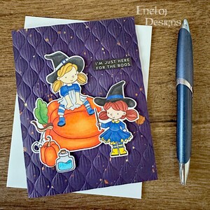 May include: A purple card with a textured background features two witches with black hats and orange pumpkins. The text on the card reads "I'm just here for the boos."