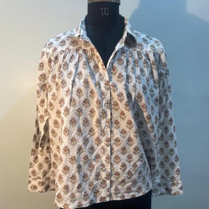 May include: A long-sleeved shirt with a white base and a repeating brown floral design. The shirt features a collared neckline and is displayed on a mannequin.