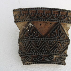 May include: A wooden printing block with intricate carved designs. The block is brown and has a black ink residue. The design features a repeating pattern of floral motifs, geometric shapes, and paisley patterns.