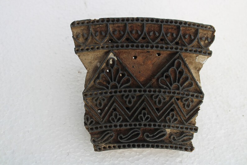 May include: A wooden printing block with intricate carved designs. The block is brown and has a black ink residue. The design features a repeating pattern of floral motifs, geometric shapes, and paisley patterns.