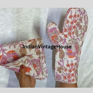 May include: A pair of pink and brown floral print oven mitts. The mitts are made of cotton and have a quilted design. The mitts are folded and displayed on a white surface. The text "IndianVintageHouse" is visible in the image.