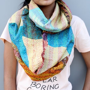 May include: A colorful silk scarf with a patchwork design in shades of blue, yellow, orange, and red. The scarf is folded into a loop and worn around the neck.