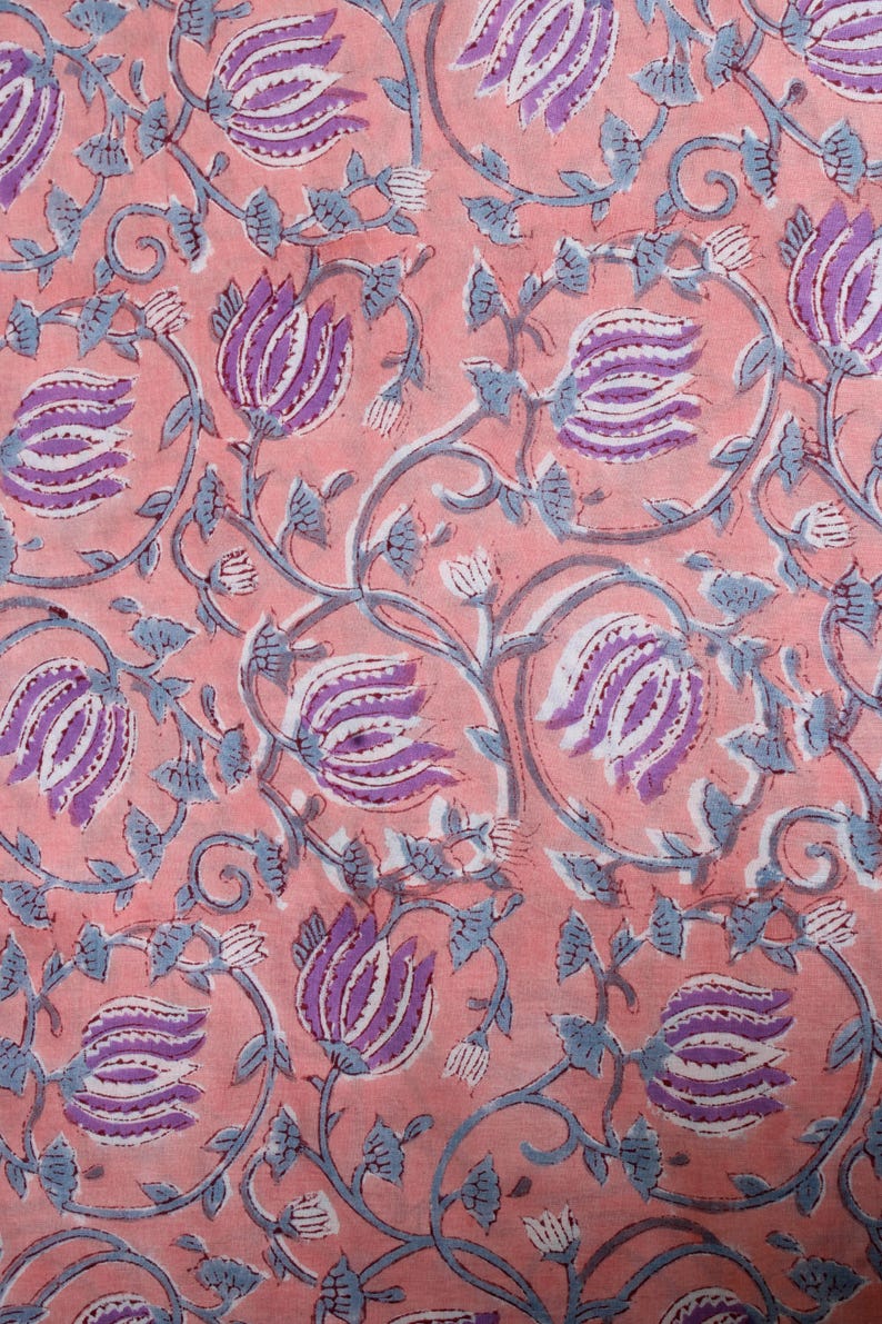May include: Close-up of a fabric with a repeating floral pattern. The design features stylized purple and white flowers with blue-grey stems and leaves on a peach-coloured background. The pattern is detailed and intricate.