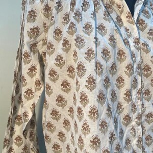 May include: A white long-sleeved shirt featuring a repeating floral pattern in brown and olive-green. The button-down shirt has a collar and cuffs, and is made of a lightweight material. The shirt's design is visible across the fabric.