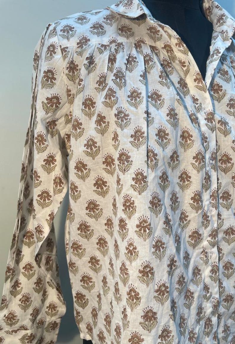 May include: A white long-sleeved shirt featuring a repeating floral pattern in brown and olive-green. The button-down shirt has a collar and cuffs, and is made of a lightweight material. The shirt's design is visible across the fabric.
