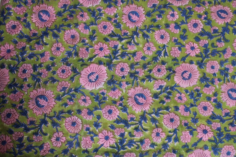 May include: A close-up of a fabric with a floral pattern. The design features pink flowers with blue centres and stems, set against a green background. The pattern is repeated across the fabric.