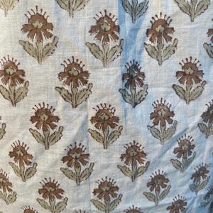 May include: Close-up of a fabric featuring a repeating floral design. The flowers are brown with light green leaves and stems, set against a light beige background. The fabric has a slightly textured appearance.