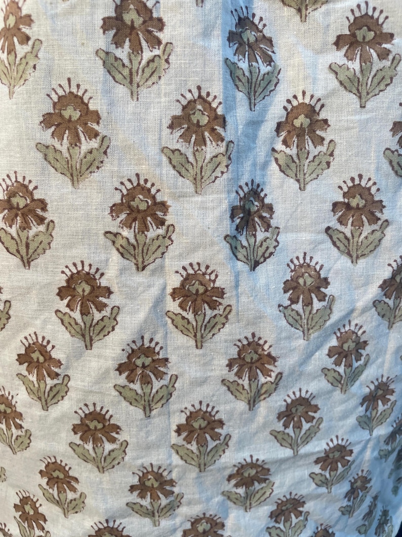 May include: Close-up of a fabric featuring a repeating floral design. The flowers are brown with light green leaves and stems, set against a light beige background. The fabric has a slightly textured appearance.
