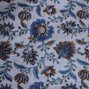 May include: Close-up of a white fabric with a floral pattern. The design features blue and brown flowers with green stems and leaves. The flowers have multiple petals and a central design, creating a detailed look.