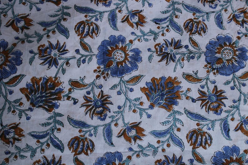 May include: Close-up of a white fabric with a floral pattern. The design features blue and brown flowers with green stems and leaves. The flowers have multiple petals and a central design, creating a detailed look.