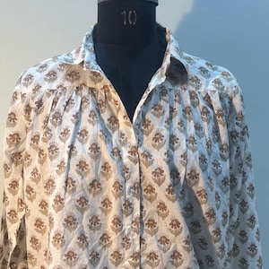 May include: A white button-down shirt with a repeating floral pattern in brown. The shirt is displayed on a black mannequin. The floral design is a stylized flower with a brown center and brown petals. The shirt has long sleeves.