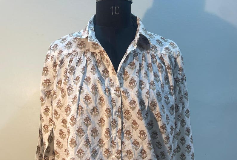 May include: A white button-down shirt with a repeating floral pattern in brown. The shirt is displayed on a black mannequin. The floral design is a stylized flower with a brown center and brown petals. The shirt has long sleeves.
