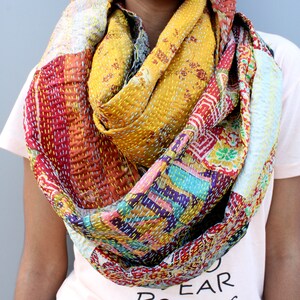 May include: A colourful patchwork scarf made of kantha stitched fabric. The scarf is a mix of red, yellow, blue, green, and white fabrics. It is folded and draped around a person's neck.