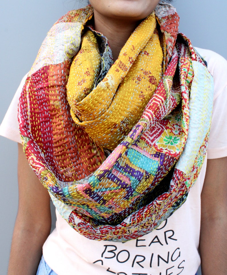 May include: A colourful patchwork scarf made of kantha stitched fabric. The scarf is a mix of red, yellow, blue, green, and white fabrics. It is folded and draped around a person's neck.