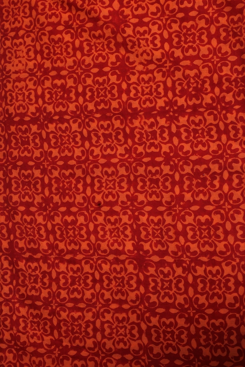 May include: Close-up of a fabric with a repeating floral pattern in shades of red and orange. The design features stylized flowers arranged in a grid-like formation, creating a textured and visually engaging surface.
