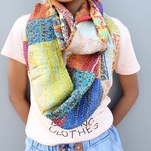 Könnte beinhalten: Ein farbenfroher Patchwork-Schal aus recycelten Saris. Der Schal ist eine Mischung aus blauen, gelben, roten und grünen Stoffen. Der Schal ist um den Hals einer Person gebunden, die ein rosa T-Shirt mit dem Aufdruck "Vintage Clothes" trägt.