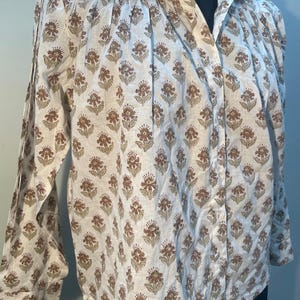 May include: A white button-down shirt with a repeating floral pattern in shades of brown and green. The long-sleeved shirt has a collared neckline and is displayed on a black mannequin.