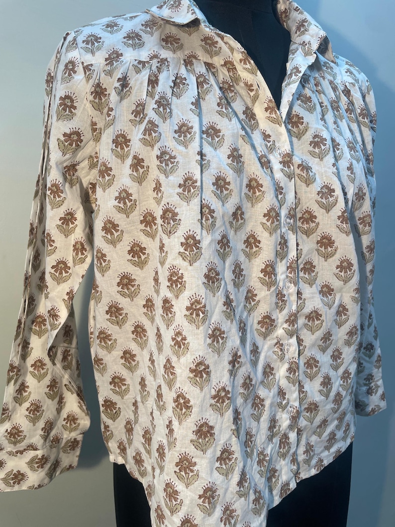 May include: A white button-down shirt with a repeating floral pattern in shades of brown and green. The long-sleeved shirt has a collared neckline and is displayed on a black mannequin.