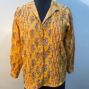 Pode incluir: Camisa de manga comprida amarelo mostarda com um padrão floral em vermelho, branco e verde. A camisa tem gola e pregas verticais na frente. O tecido parece leve.